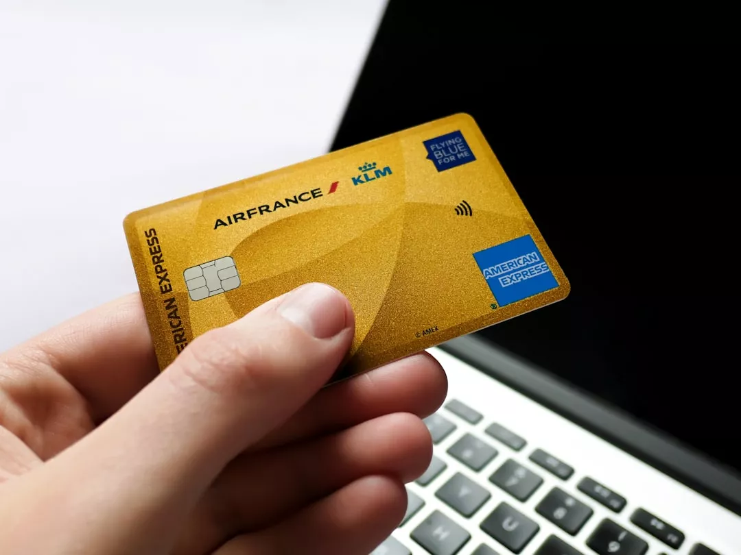 Hand holding a gold american express card near laptop.